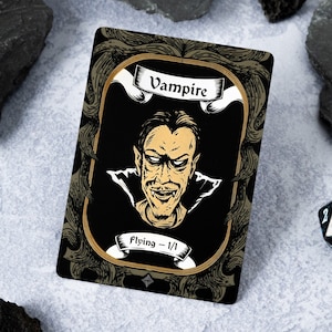 May include: A black and gold card with an illustration of a vampire with a white banner that says "Vampire" and a white banner at the bottom that says "Flying - 1/1".