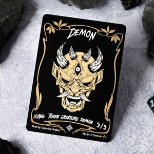 Könnte beinhalten: Eine schwarze Karte mit goldenen Akzenten, die eine Dämonenillustration zeigt. Die Karte lautet "DEMON FLYING TOKEN CREATURE DEMON 5/5 Made by Legendary Artifacts Flip for 5 Demons".