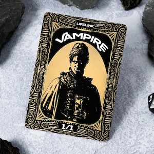 May include: A black and gold card game card with the text "Lifelink Vampire" and a black and white illustration of a vampire. The card has a 1/1 power and toughness.