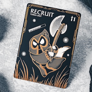 Puede incluir: Una tarjeta negra y dorada de la Unidad de Fichas de Reclutamiento con una figura de dibujos animados con un hacha. La tarjeta tiene el número 1 en la esquina superior derecha y el texto "RECRUIT TOKEN UNIT" en la parte superior. La tarjeta tiene un borde dorado y detalles dorados.