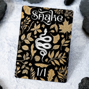 May include: A black card with a gold floral pattern and a white snake illustration. The card has the word "Snake" in gold lettering and the number "1/1" in white lettering.