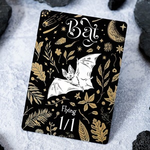 May include: A black and gold card featuring a white bat illustration. The card has a black background with gold floral designs and the text "Bat" in a decorative font. The card also has the text "Flying 1/1".