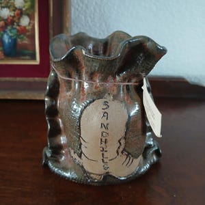 May include: A ceramic vase with a ruffled top and a textured, mottled brown and tan finish. The vase has a light-colored oval with the word "SANDHILLS" written vertically. A small tag hangs from the rim.