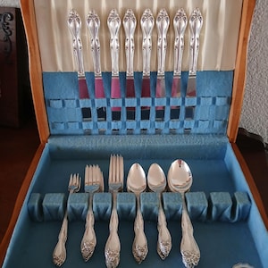 May include: A wooden silverware case with a blue velvet interior holds a set of silver utensils. The set includes knives, forks, and spoons, all with ornate handles. The case is open, revealing the arrangement of the silverware.