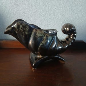 May include: A dark, glossy ceramic vase in the shape of a cornucopia. The vase has a curved, horn-like shape with a flared opening and a decorative, curled end. The color is a mix of dark brown and black with hints of green.