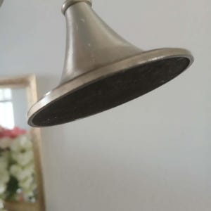 May include: Close-up of a brushed nickel-finished shower head. The shower head has a conical shape with a flat, dark-colored underside. The fixture is mounted on a curved arm, and the background is a plain white wall.