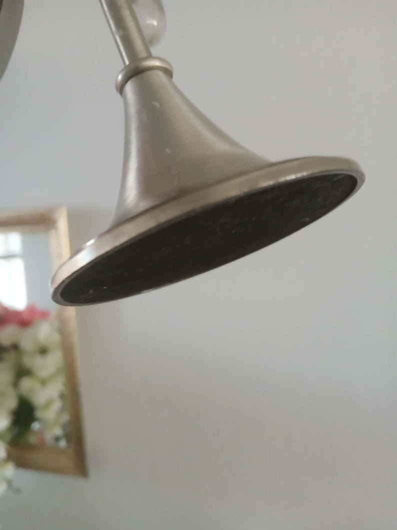 May include: Close-up of a brushed nickel-finished shower head. The shower head has a conical shape with a flat, dark-colored underside. The fixture is mounted on a curved arm, and the background is a plain white wall.