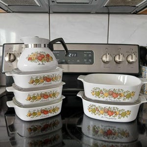 May include: A set of vintage Pyrex casserole dishes with floral designs and the text "Le Creuset" on the bottom. The dishes are stacked on top of each other and are sitting on a black stovetop.