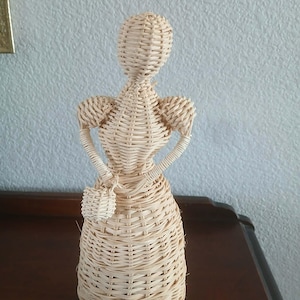 May include: A woven wicker figurine of a woman in a dress, holding a small purse. The figure is made of light brown wicker and has a detailed woven pattern. The figurine is standing on a dark brown surface.