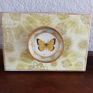 May include: A framed artwork with a yellow butterfly on a light blue background. The frame is decorated with gold accents and the background is a light green with a floral pattern.