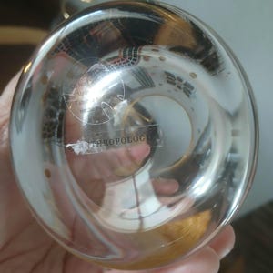 May include: Clear glass Anthropologie decorative object with a round shape. The glass has a slight amber tint and is partially transparent. The bottom of the object has a sticker with text. The object is held in a hand.