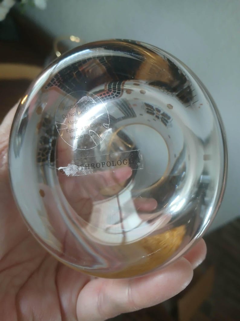 May include: Clear glass Anthropologie decorative object with a round shape. The glass has a slight amber tint and is partially transparent. The bottom of the object has a sticker with text. The object is held in a hand.