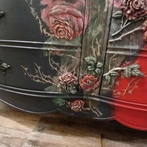 May include: A black and red painted dresser with rose and thorn details. The roses are in relief, with a mix of colors including pink, red, and green. The dresser has a curved front and decorative hardware.