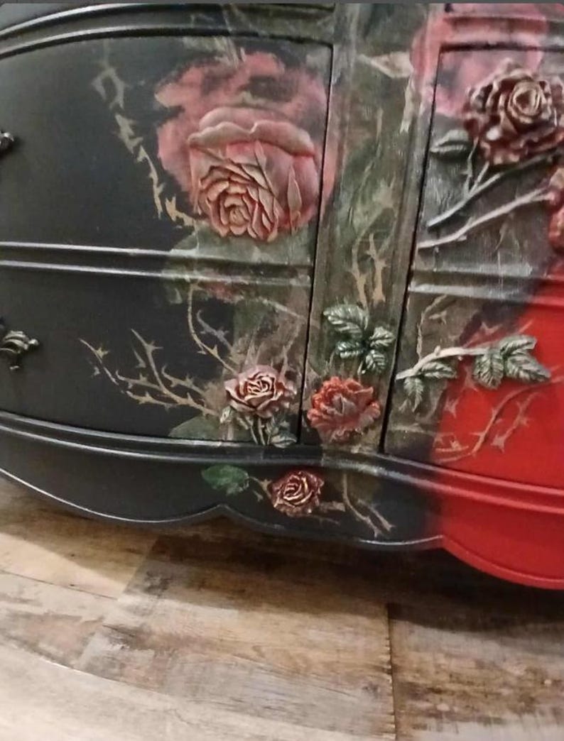 May include: A black and red painted dresser with rose and thorn details. The roses are in relief, with a mix of colors including pink, red, and green. The dresser has a curved front and decorative hardware.