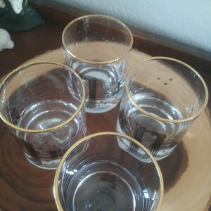 May include: Four clear drinking glasses with gold rims and a black and gold geometric design. The glasses are arranged on a wooden serving tray. These glasses are perfect for serving drinks. Ideal for a home bar.