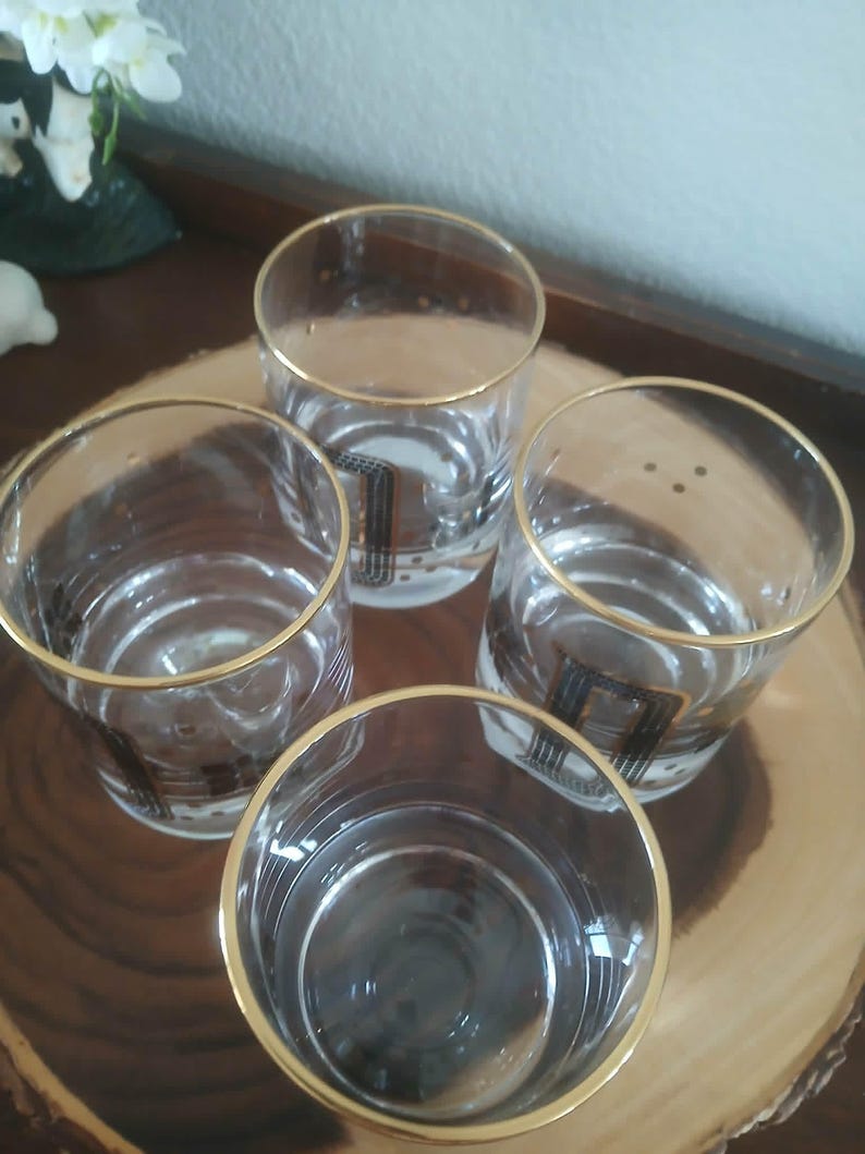 May include: Four clear drinking glasses with gold rims and a black and gold geometric design. The glasses are arranged on a wooden serving tray. These glasses are perfect for serving drinks. Ideal for a home bar.