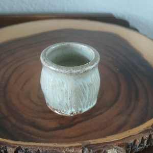 May include: A small, handmade ceramic pot with a light beige glaze and vertical white streaks. The pot has a rounded shape and a slightly textured surface. It sits on a dark wooden surface.
