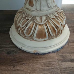 May include: A close-up of the base of a decorative pedestal. The base is off-white with gold accents and a floral design. The pedestal sits on a round, off-white base.