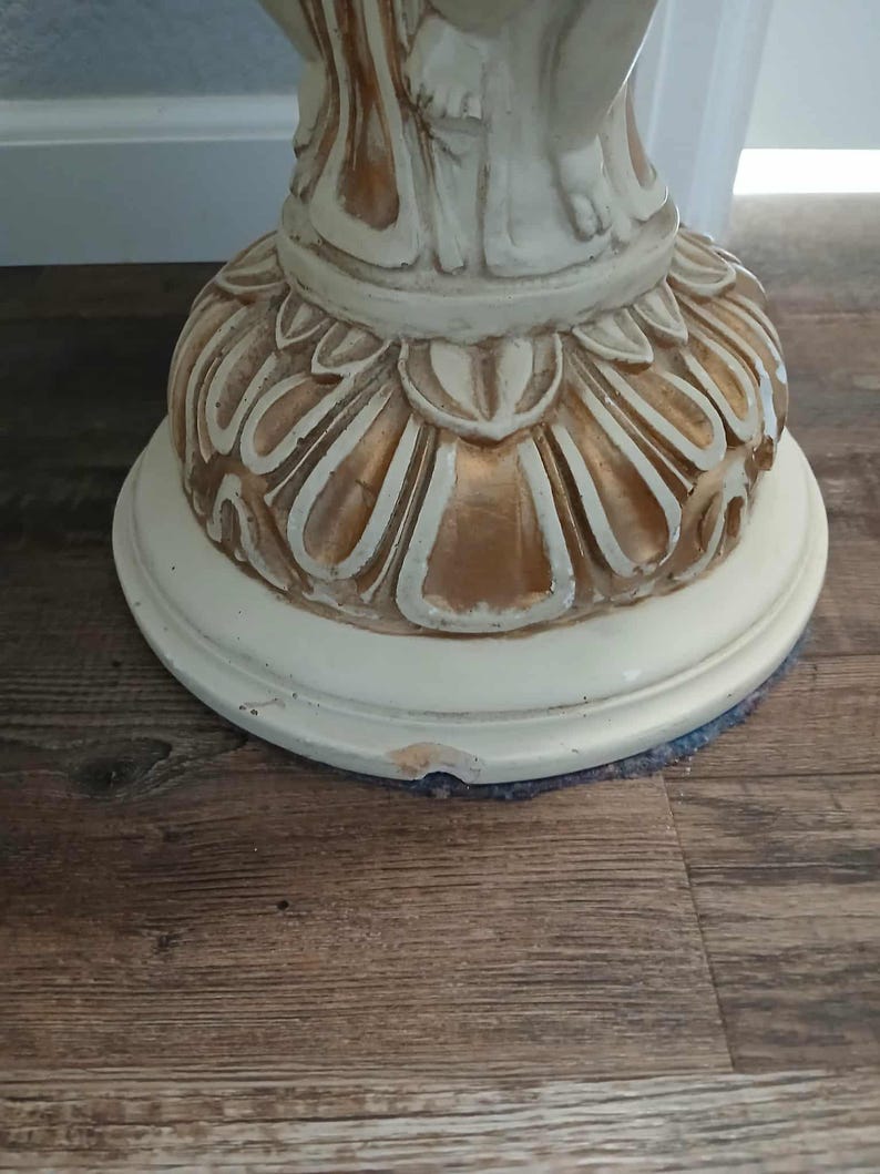 May include: A close-up of the base of a decorative pedestal. The base is off-white with gold accents and a floral design. The pedestal sits on a round, off-white base.