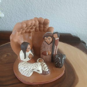 May include: A ceramic nativity scene featuring figures of Mary, Joseph, baby Jesus, a lamb, and a sheep. The figures are set against a backdrop of a large hand. The color palette includes shades of brown, white, and black.