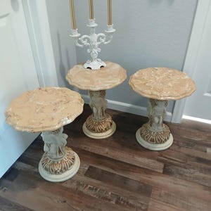 May include: Three decorative side tables with round, textured tops in shades of beige and cream. The bases feature cherub figures and ornate detailing. A white candelabra with gold candles sits on one table. The tables are on a wood floor.