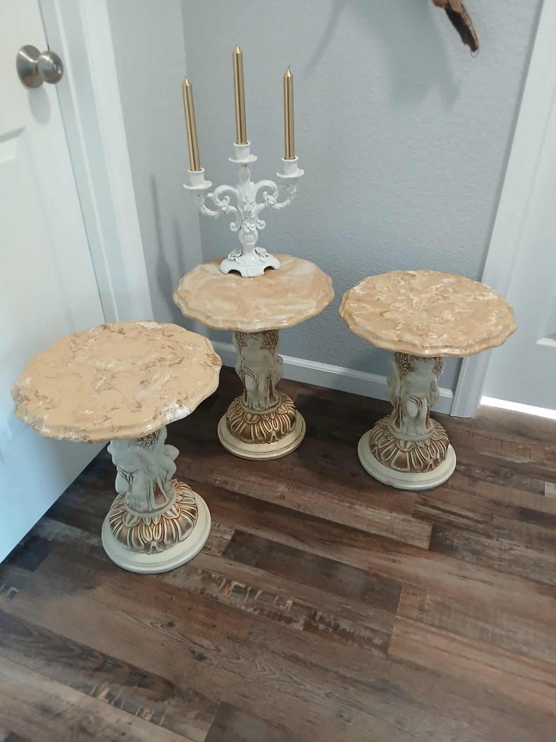 May include: Three decorative side tables with round, textured tops in shades of beige and cream. The bases feature cherub figures and ornate detailing. A white candelabra with gold candles sits on one table. The tables are on a wood floor.