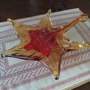 May include: A star-shaped glass dish with a red center and amber-colored edges. The dish is made of blown glass and has a unique, organic shape.