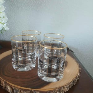 May include: Four clear drinking glasses with gold rims and a black and gold geometric design. The glasses are arranged on a round wooden serving tray with a natural edge. The glasses are on a dark brown table.