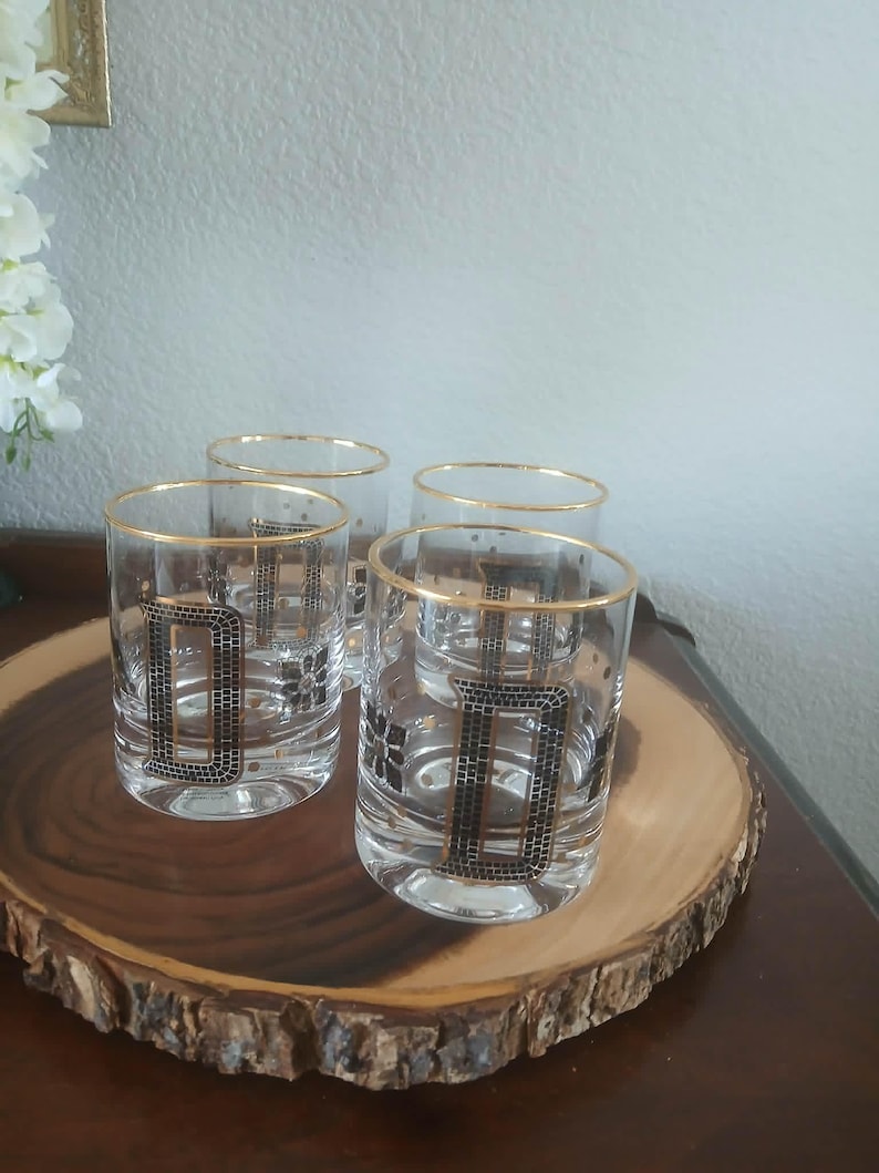 May include: Four clear drinking glasses with gold rims and a black and gold geometric design. The glasses are arranged on a round wooden serving tray with a natural edge. The glasses are on a dark brown table.