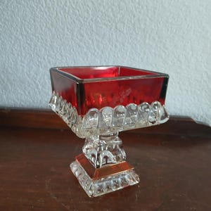 May include: A vintage red glass pedestal bowl with a square top and a clear, ornate base. The bowl has a ruby red hue, and the base features a detailed, geometric design. The bowl is likely used for decorative purposes.
