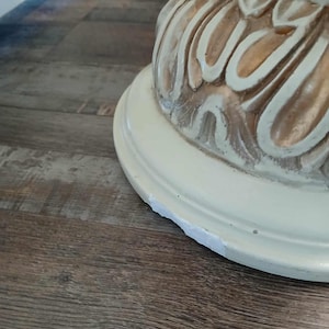May include: Close-up of a cream-colored, ornate table base with carved floral details. The base has a round, slightly chipped bottom. The wood floor has a brown, wood grain pattern.