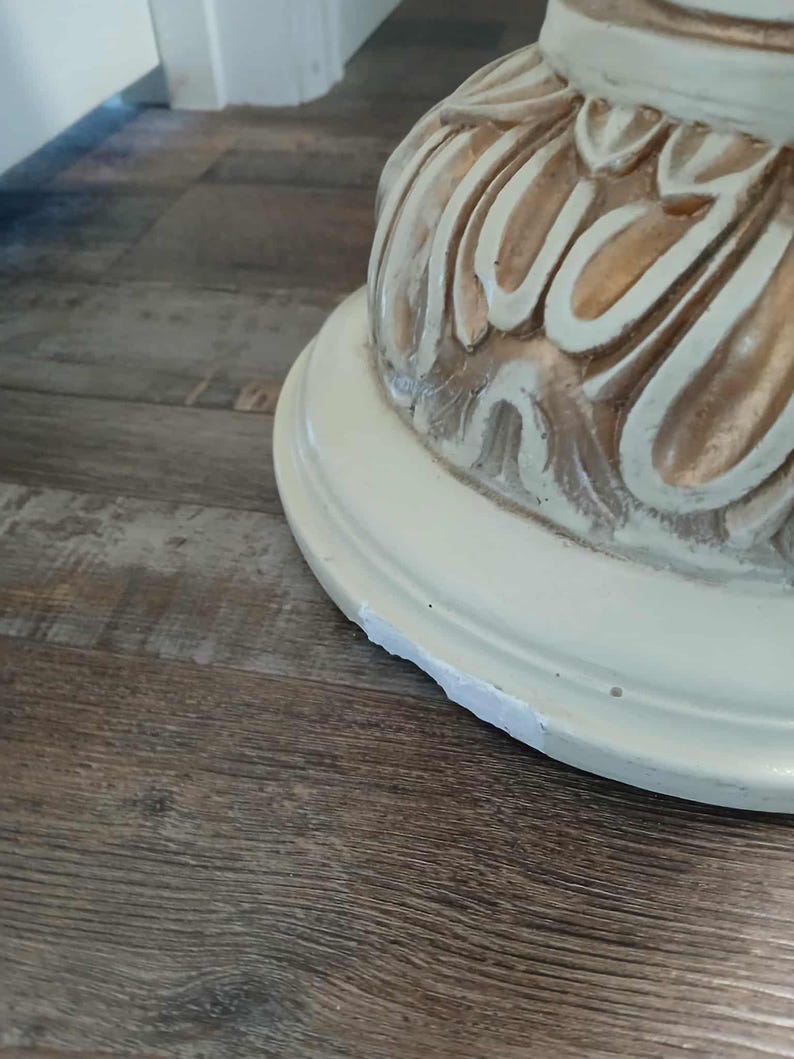 May include: Close-up of a cream-colored, ornate table base with carved floral details. The base has a round, slightly chipped bottom. The wood floor has a brown, wood grain pattern.