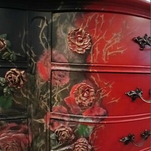 May include: A painted dresser with a striking two-tone design. One side is black with red roses and the other is red with a floral and thorn motif. The drawers feature ornate, dark metal handles and raised rose details.