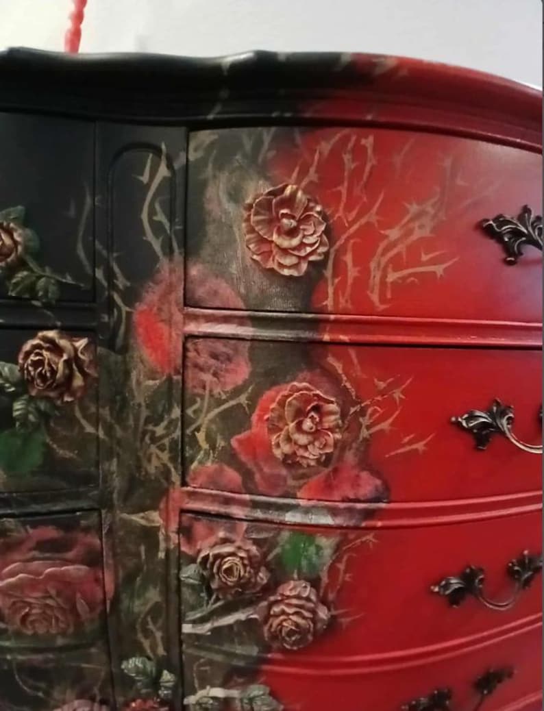 May include: A painted dresser with a striking two-tone design. One side is black with red roses and the other is red with a floral and thorn motif. The drawers feature ornate, dark metal handles and raised rose details.