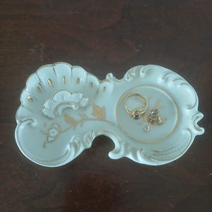 May include: A light blue and white porcelain ring dish with gold accents. The dish has a floral design and holds two rings. The dish is approximately 6 inches long and 4 inches wide.