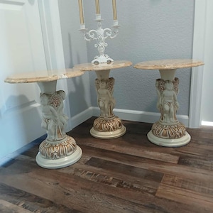 May include: Three decorative side tables with marble-look tops. Each table has a cherub-adorned pedestal base in cream and gold tones. A white candelabra with three candles sits on one table. The tables are on a wood-look floor.