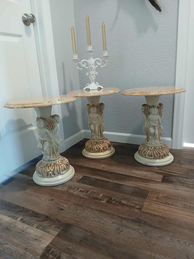 May include: Three decorative side tables with marble-look tops. Each table has a cherub-adorned pedestal base in cream and gold tones. A white candelabra with three candles sits on one table. The tables are on a wood-look floor.