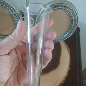 May include: A clear glass bud vase with a ruffled top. The vase is held in a hand, with a blurred background of a silver picture frame and a wooden slice.