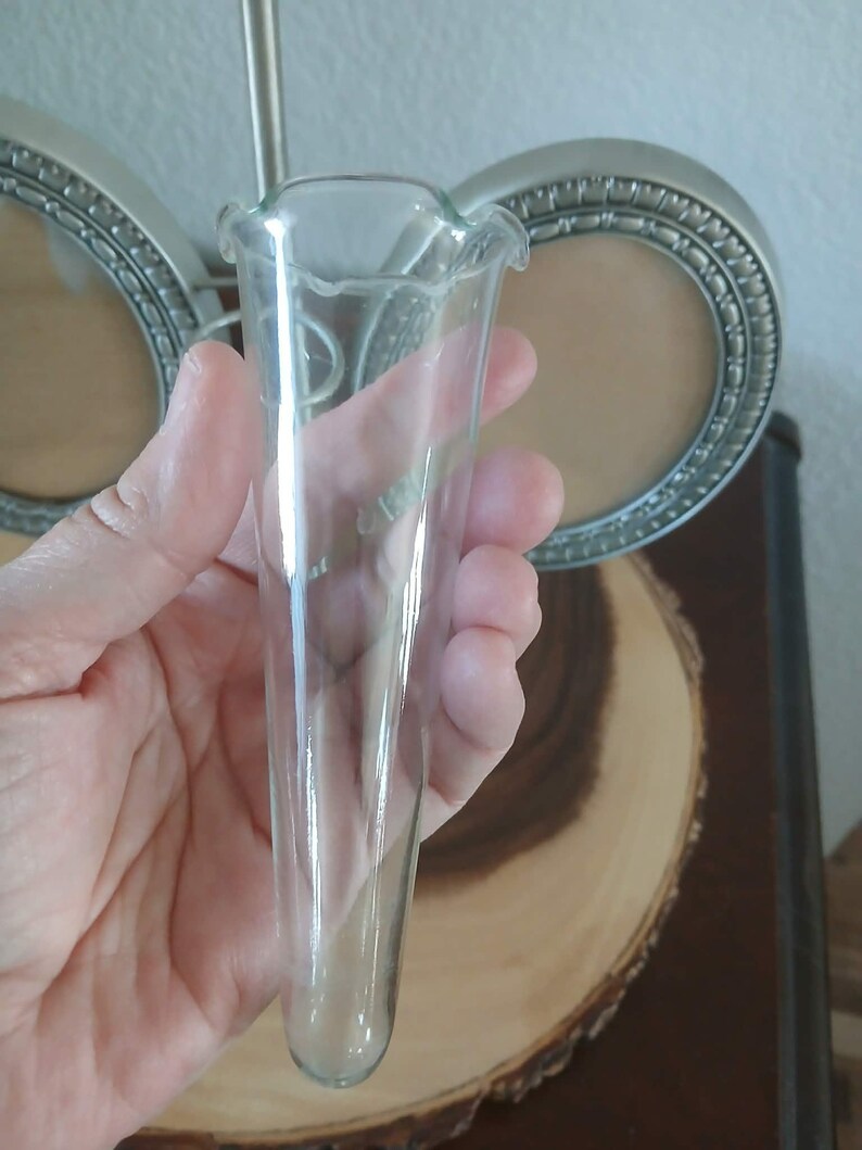 May include: A clear glass bud vase with a ruffled top. The vase is held in a hand, with a blurred background of a silver picture frame and a wooden slice.