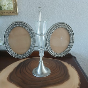 May include: A silver-toned metal photo frame with two oval openings and a central vase. The frames have a decorative border. The vase is clear glass. The item is displayed on a wooden slice.