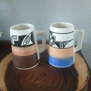 May include: Two ceramic mugs with white handles and a geometric design in black. The mugs have a color gradient from brown to tan and blue. They are displayed on a wooden slab.