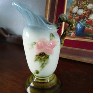 May include: A vintage ceramic pitcher with a light blue spout and a white body adorned with a pink floral design. The handle is a dark green, and the base is a darker green with brown stripes. The pitcher is likely for decorative use.