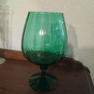 May include: A large, emerald green glass goblet with a wide bowl and a stemmed base. The glass has a rippled texture and a scalloped rim. The goblet is sitting on a dark wooden surface.