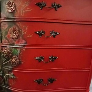 May include: Red painted dresser with four drawers and ornate black handles. The left side features a floral design with raised rose details. The dresser has a curved front and legs, creating a vintage aesthetic.
