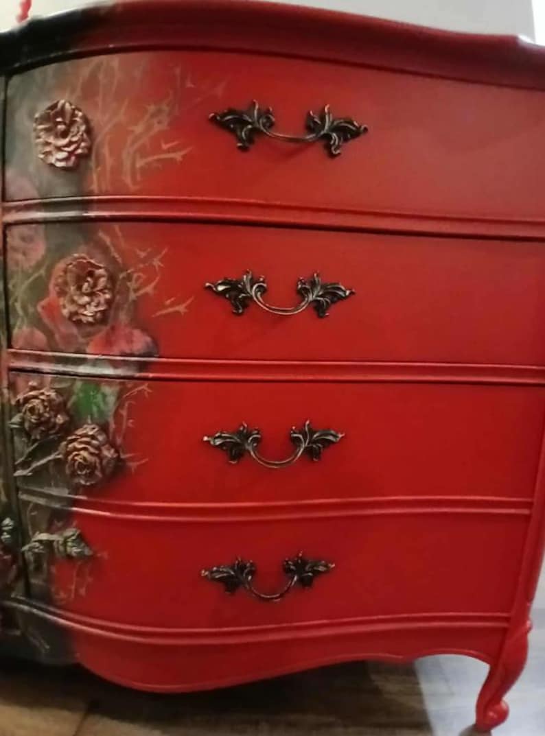 May include: Red painted dresser with four drawers and ornate black handles. The left side features a floral design with raised rose details. The dresser has a curved front and legs, creating a vintage aesthetic.