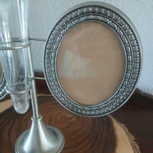 May include: A silver-toned metal photo frame with an oval opening and decorative border. The frame is part of a decorative stand that also holds a small glass vase. The stand has a round base and a slender stem.