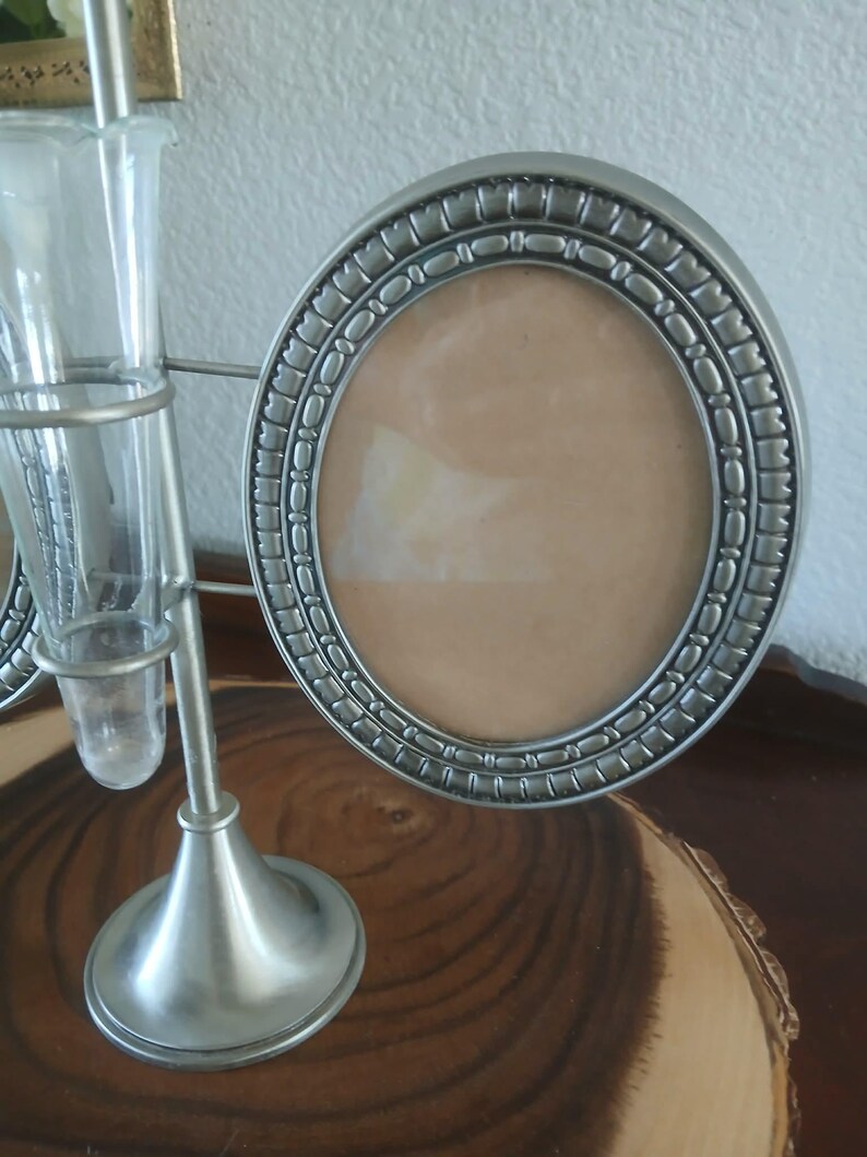 May include: A silver-toned metal photo frame with an oval opening and decorative border. The frame is part of a decorative stand that also holds a small glass vase. The stand has a round base and a slender stem.
