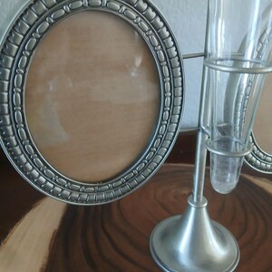 May include: A silver-toned metal oval picture frame with a decorative border sits next to a silver-toned metal bud vase holder with two glass vases. The frame has a light brown backing.
