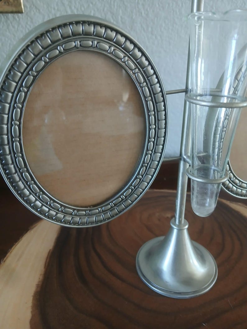 May include: A silver-toned metal oval picture frame with a decorative border sits next to a silver-toned metal bud vase holder with two glass vases. The frame has a light brown backing.