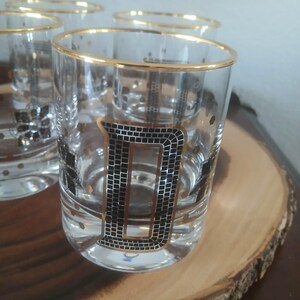 May include: A set of six vintage clear glass tumblers with gold rims and a black letter "D" design. The glasses have a mid-century modern aesthetic and are displayed on a wooden serving tray. Each glass has a capacity of approximately 240 ml.